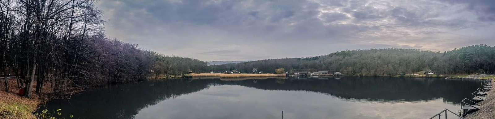 A still lake below forested hills, with houses and a windmill along the far shore, under a wide sky. ASTRA Park, Sibiu.